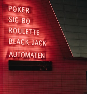 Red neon letters are mounted on a dark tiled wall, displaying the words Poker, Sic Bo, Roulette, Black Jack, and Automaten.