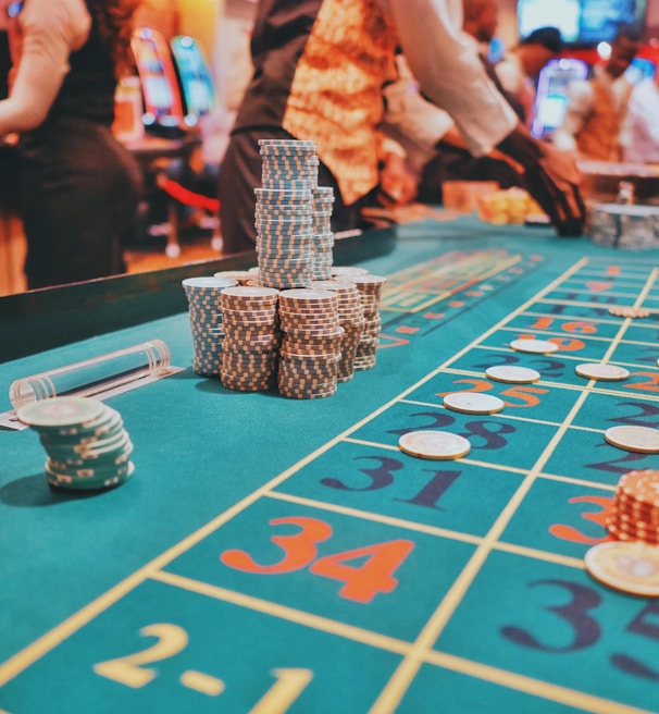 A casino scene with a roulette table features prominently, covered in green felt with colorful numbers and betting squares. Several stacks of poker chips are placed on different numbers, and the hands of several people are visible as they interact with the table. In the background, there are slot machines and more people, creating a vibrant and busy atmosphere.