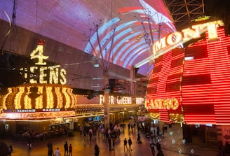A bustling nighttime scene with illuminated casino signage towering over a busy pedestrian street. Neon lights in red and yellow highlight the names of various establishments. People are walking in different directions, enjoying the vibrant atmosphere.