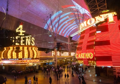 A bustling nighttime scene with illuminated casino signage towering over a busy pedestrian street. Neon lights in red and yellow highlight the names of various establishments. People are walking in different directions, enjoying the vibrant atmosphere.