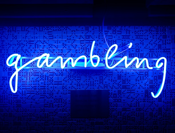A bright blue neon sign spells out the word 'gambling' in cursive script. The background features a textured wall covered in domino tiles, also illuminated by the blue light, creating a vivid and visually striking scene.