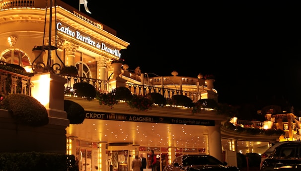 A luxurious casino at night, illuminated with warm, golden lights. The building features elegant architecture with columns and decorative elements. A few people are walking towards the entrance, and there are a couple of parked cars nearby.