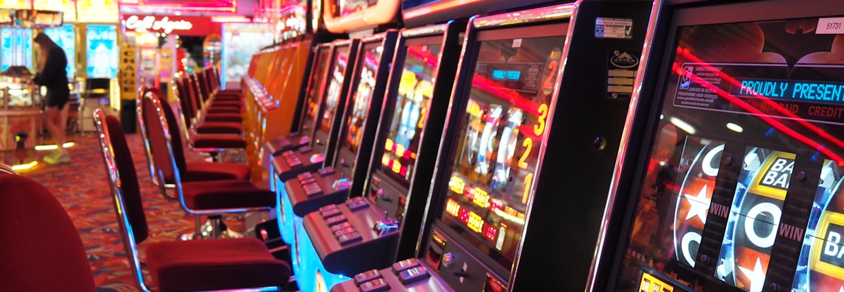 A brightly lit casino interior featuring a row of colorful slot machines. The neon lights create a vibrant atmosphere, with patterned carpeting adding to the decor. Stools are aligned neatly in front of each machine.
