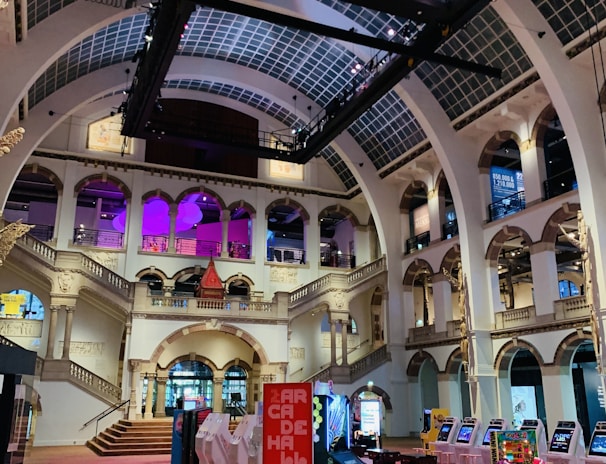 The interior of a large, ornately designed building with arched ceilings and intricate architectural details. The space features gaming machines lined up along the floor, under a high ceiling with a grid pattern. Lighting in purple and white tones enhances the ambiance.