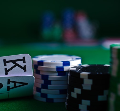 A casino scene featuring two playing cards, the ace and king of clubs, partially concealed by a player's hand. Nearby, there are stacks of multicolored poker chips on a green felt table, with blurred colorful background elements suggesting other chip stacks or cards.