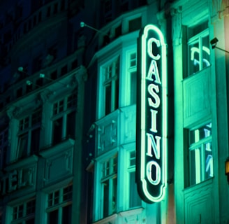 A vibrant neon sign displaying the word 'CASINO' is prominently illuminated against the facade of a building, showcasing intricate architectural details and a classic design. The sign emits a bright teal glow, casting light on the surrounding area and creating a lively atmosphere at night.