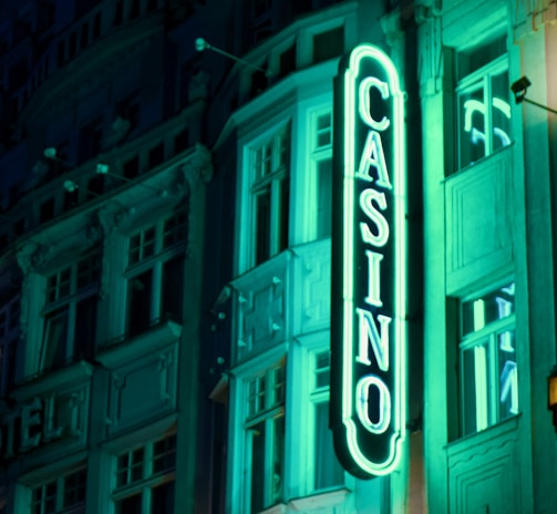 A vibrant neon sign displaying the word 'CASINO' is prominently illuminated against the facade of a building, showcasing intricate architectural details and a classic design. The sign emits a bright teal glow, casting light on the surrounding area and creating a lively atmosphere at night.