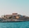 A seaside view of a large building labeled as a casino, situated on a rocky shore. The building is surrounded by palm trees and other smaller structures. The calm sea water reflects the sky, and there are several closed umbrellas lined along the waterfront.
