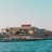 A seaside view of a large building labeled as a casino, situated on a rocky shore. The building is surrounded by palm trees and other smaller structures. The calm sea water reflects the sky, and there are several closed umbrellas lined along the waterfront.