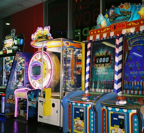 A row of brightly colored arcade game machines is arranged in a line against the wall. Each machine features vibrant lights and designs, with themes ranging from claw games to game show-inspired setups. The area has a playful and inviting atmosphere, suitable for entertainment or amusement centers.