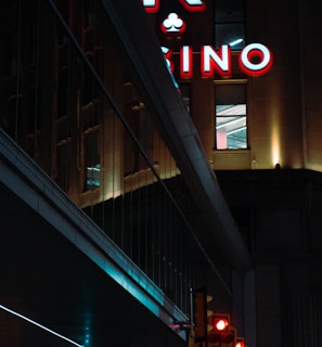 A night scene featuring a building with illuminated neon signage displaying 'CASINO'. The environment is urban with reflections on glass windows and glowing street traffic lights below. The atmosphere is dimly lit, with artificial lighting casting vibrant colors.