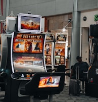 A collection of slot machines is situated inside a building with bright, colorful screens displaying various games and gaming themes. A person is seated nearby, seemingly relaxed with luggage beside them, and the atmosphere is lively with electronic displays.