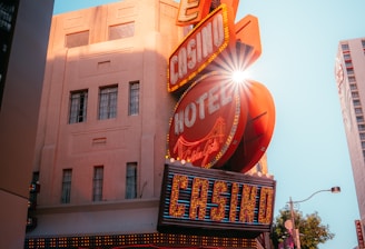 A vibrant city scene featuring a tall, illuminated casino sign with retro-style neon lights. The sign dominates the building facade with the words 'CASINO' and 'HOTEL'. Sunlight peeks through, creating a dramatic lens flare effect. The street is lively, with pedestrians casually walking and a hint of urban hustle.