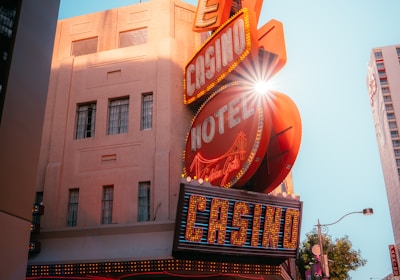 A vibrant city scene featuring a tall, illuminated casino sign with retro-style neon lights. The sign dominates the building facade with the words 'CASINO' and 'HOTEL'. Sunlight peeks through, creating a dramatic lens flare effect. The street is lively, with pedestrians casually walking and a hint of urban hustle.